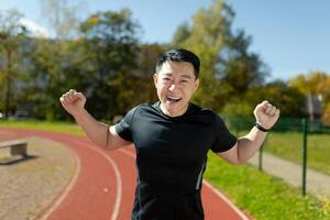 un joven asiático hombre, un atleta, un corredor celebra un victoria en un carrera, en un maratón. el contento hombre es en pie en el estadio en el corriendo pista, elevado su manos arriba, se regocija en el cámara. foto