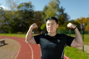 Portrait of a young Asian man, a sportsman, a bodybuilder standing in a stadium and showing a gesture of strength with his hands and muscles. Look seriously into the camera. photo