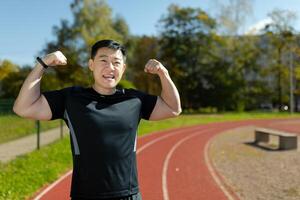 retrato de un asiático masculino deportista en pie en un estadio y demostración el cámara un poder gesto con su manos, demostración apagado su tonificado músculos y cuerpo. foto