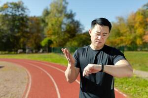 retrato de un joven asiático masculino atleta, un corredor en pie en un rueda de andar en un estadio y comprobación el hora en un inteligente mirar. insatisfecho con el resultado, él se extiende su manos en frustración. foto