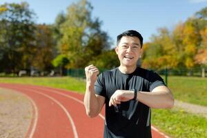 retrato de un joven asiático hombre haciendo Deportes y corriendo en el estadio, mirando a el inteligente reloj y contento acerca de el bueno resultado. sonriente a el cámara, él muestra un victorioso gesto con su mano. foto