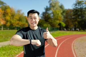 A young Asian man, an athlete standing on a running track in a stadium, checking at the time on a smart watch, pointing a super finger. He smiles, satisfied with the result of the race, marathon. photo