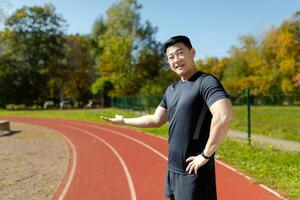 Young Asian athlete, coach standing in stadium and pointing with hands to running track. Invites to training, presents and popularizes sports. photo