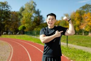 Portrait of Asian young male athlete standing on stadium field in black sportswear and pointing finger at arm muscles, smiling at camera. photo