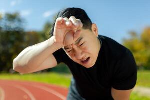 Close-up of sportsman having severe headache and dizziness, asian man in stadium doing fitness and active exercise, male runner in sportswear overworked by training on sunny day. photo