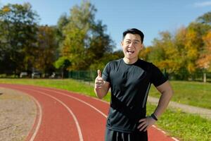 Portrait of a smiling young Asian man standing in a stadium in sportswear and pointing super finger at the camera. photo