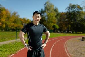 Portrait of a smiling and confident Asian male athlete standing in front of the camera in a stadium with his arms at his sides. photo