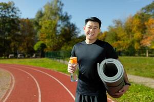 Portrait of a young Asian male athlete, trainer standing in a stadium, holding an exercise and yoga mat and a bottle of water. Smiling at the camera. photo