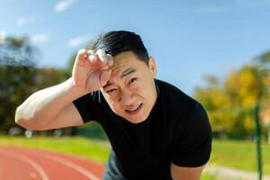 Close-up photo. Portrait of a young Asian male sportsman tiredly bent over at the stadium, resting after jogging and training, looking at the camera. photo