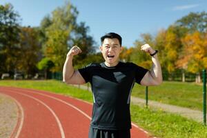 Portrait of a happy young Asian male athlete standing in a stadium, rejoicing at the camera for winning and showing the camera a gesture of strength and victory with his hands. photo