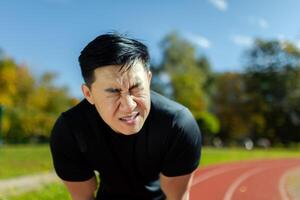 Close-up of sportsman having severe headache and dizziness, asian man in stadium doing fitness and active exercise, male runner in sportswear overworked by training on sunny day. photo