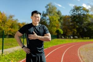 Portrait of a young man Asian sportsman, athlete, runner standing in the stadium and holding his chest with his hand. Feels pain, attack, exhaustion, rests. photo