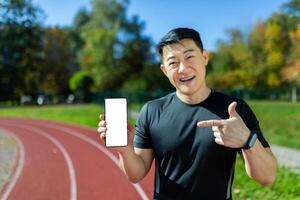 Successful and satisfied Asian athlete on training smiling and looking at camera man showing smartphone screen to camera with blank space on white background, athlete training on sunny day at stadium photo
