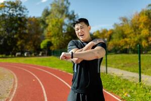 asiático deportista estiramientos su hombro, hombre en el estadio después corriendo y activo físico ejercicios estiramientos su brazo articulaciones, tiene hombro dolor, brazo músculo espasmo. foto