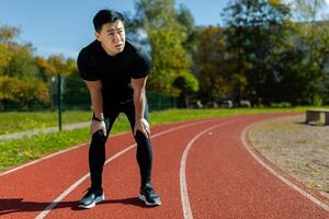 Asian sportsman tired after running and active physical exercise, man taking breath and rest on sunny day in stadium, overtired after running. photo