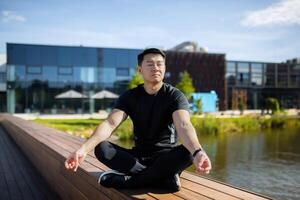 asiático hombre medita en el loto posición, hombre en el parque cerca el lago realiza yoga ejercicios, deportista descansa con un cerrado barbilla después activo físico ejercicios y correr. foto