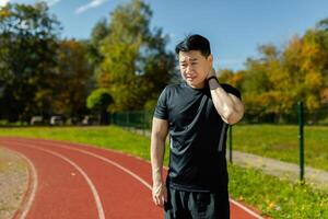 Portrait of a young Asian male athlete standing in the stadium and holding his neck, pulled a muscle, feels severe pain, massages, rests. photo