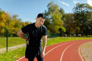 Asian sportsman has severe pain in back and abdominal muscles, man peed training in stadium on sunny day massaging side of back, athlete on jogging pulled muscle. photo