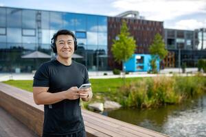 A young Asian male athlete in headphones and with a phone is standing outside and checking his exercise, running, marathon results. Installs a sports app. photo