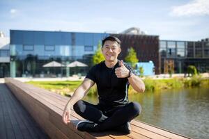 Portrait of a young Asian sportsman man sitting on a bench by the water in the lotus yoga position, resting, smiling and pointing super finger at the camera. photo