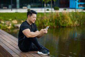 A young Asian sportsman man is sitting resting on a wooden bench near the lake and using a mobile phone. photo