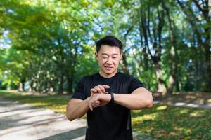 sonriente y exitoso asiático hombre trotar y hacer ejercicio en el parque, hombre mirando a inteligente reloj y comparando resultados, atleta contento con resultados y sonriente. foto