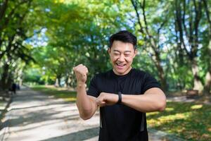 Marathon. A young Asian athlete man stands in the park in sports clothes, looks at a fitness bracelet, a smart watch, rejoices at the result of a run, shows a winning gesture with his hand. photo