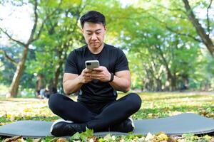 joven hermoso masculino asiático deportista se sienta en el parque en un estera en negro ropa de deporte y usos un móvil teléfono. descansando después activo capacitación. foto