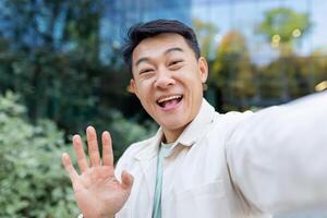 Asian programmer talking on call using app on phone, view camera man waving and talking cheerfully, outside office building on break. photo