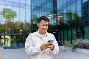 Asian man using phone outside office building, freelancer businessman typing message and smiling, programmer in casual clothes browsing online web pages from app. photo