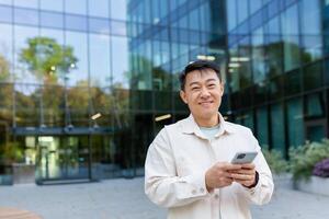 Portrait of successful Asian businessman in casual clothes, man with phone in hands smiling and looking at camera, business owner outside office building, satisfied freelancer startup entrepreneur. photo