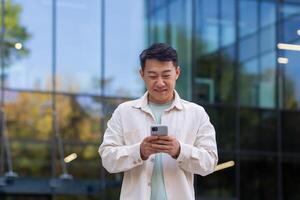 Portrait of young Asian man designer, freelancer, businessman standing near office center and using phone, reading news, writing message, checking mail, waiting for meeting. photo