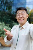 Asian programmer talking on call using app on phone, view camera man waving and talking cheerfully, outside office building on break photo