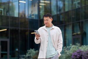 exitoso asiático hombre en casual ropa usos el teléfono, hombre camina en el ciudad cerca un moderno edificio, diales un mensaje, llamadas, empresario después trabajar. foto