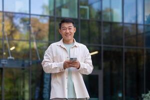 Portrait of happy and successful asian man with phone, man smiling and looking at camera, programmer businessman using app on smartphone to browse internet pages, walking outside office building. photo