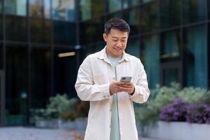 Asian man using phone outside office building, freelancer businessman typing message and smiling, programmer in casual clothes browsing online web pages from app. photo