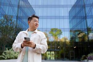 Smiling asian man in casual shirt walking in city using app on smartphone, man looking to side on background of office building from outside. photo