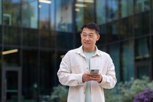 Smiling asian man in casual shirt walking in city using app on smartphone, man looking to side on background of office building from outside. photo
