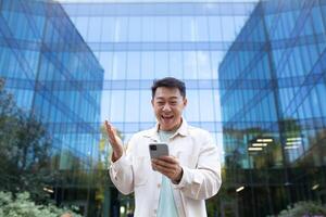 retrato de un joven asiático hombre en pie cerca un vaso rascacielos y participación un teléfono. contento recibido bueno noticias, lee mensajes, chats en llamar. foto