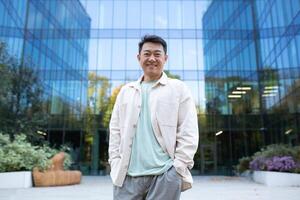 Portrait of a young handsome successful Asian male programmer, freelancer, student. Standing in front of office center outside, hands in pockets, smiling and looking at camera. photo