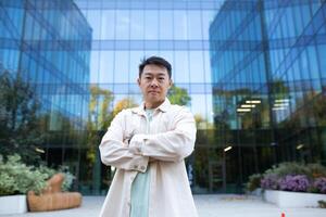 Portrait of a serious young handsome successful male Asian programmer, freelancer. Standing in front of the office center outside, arms crossed, looking at the camera. photo