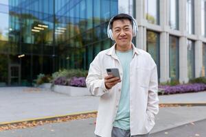 Portrait of asian man outdoors with phone and headphones for listening to music, happy man using smartphone app smiling and looking at camera. photo