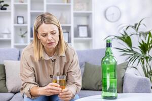 An alcoholic woman drinks strong alcohol, is depressed and sits alone at home on the sofa in the living room. photo