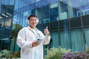 A young male Asian student stands outside the campus, co-working space. He listens to music, a podcast, an audiobook in white headphones from the phone, points a super finger at the camera, smiles. photo