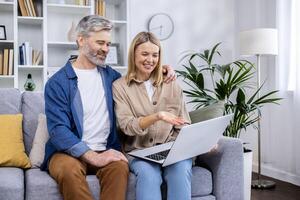 Senior mature couple man and woman sitting at home in living room on sofa, people in love using laptop, having fun choosing products in online internet store together. photo