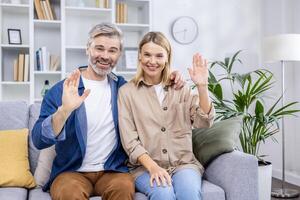 Happy family couple mature man and woman sitting together on sofa in living room and exchange looking at camera smiling and waving hand greeting gesture, call webcam view. photo