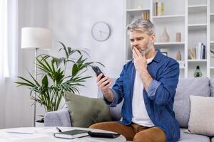 Mature adult man upset and confused reading bad news online from phone, gray haired person sitting on sofa in living room at home working with documents home finances and budget on paper work. photo