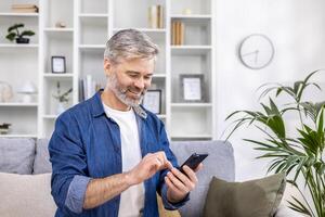 Mature adult man close-up at home smiling and using phone in living room sitting on sofa, person typing message and browsing web page. photo