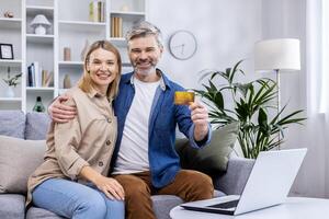 Portrait of happy family at home, adult mature couple man and woman sitting on sofa in living room and looking cheerfully at camera, man holding bank credit card in hands, happy with online shopping. photo