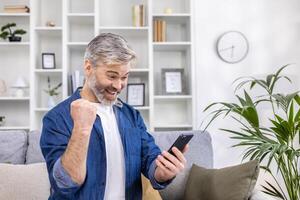 Mature gray haired man alone at home using phone and celebrating victory success happy holding hand up, adult person sitting on sofa in living room on sunny day. photo